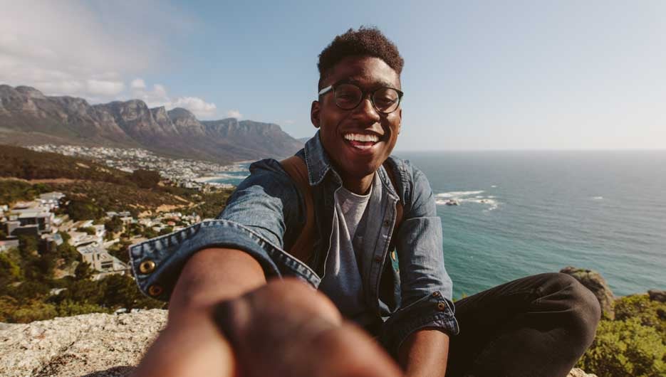 Smiling african guy sitting on top of a mountain taking selfie against seascape. Man enjoying his holiday and making self portrait against beautiful landscape.